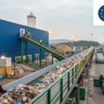 Industrial waste processing facility with a long trash conveyor overlooking a blue building; workers in safety vests on the stairs and near the conveyor.